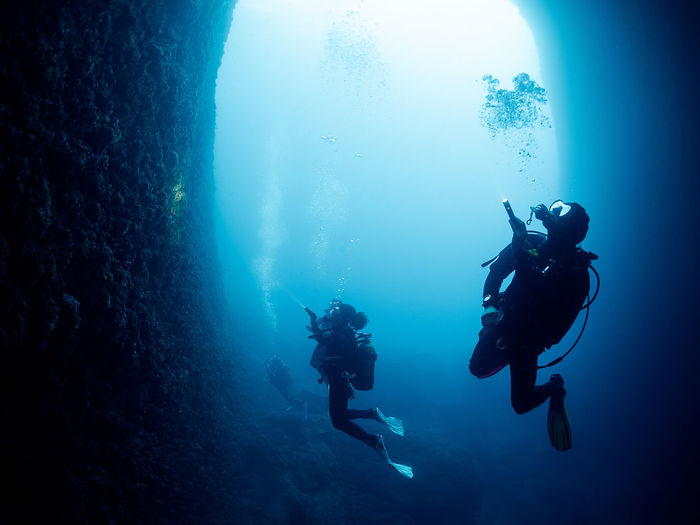 Unrecognizable professional divers with scuba and flippers swimming in narrow dark cave with rough stony formations undersea in deep clear water