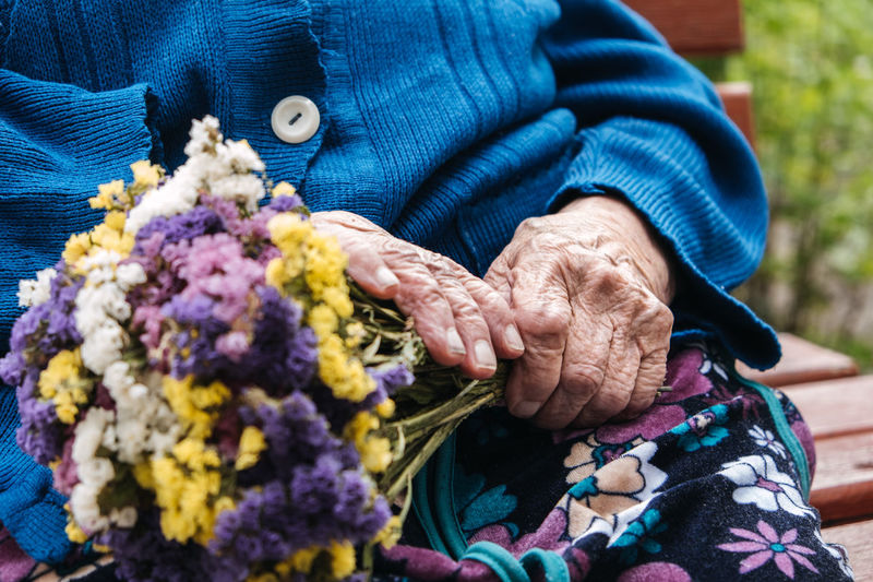 Senior elderly woman with gift, flowers bouquet and basket of groceries sits on bench outdoor