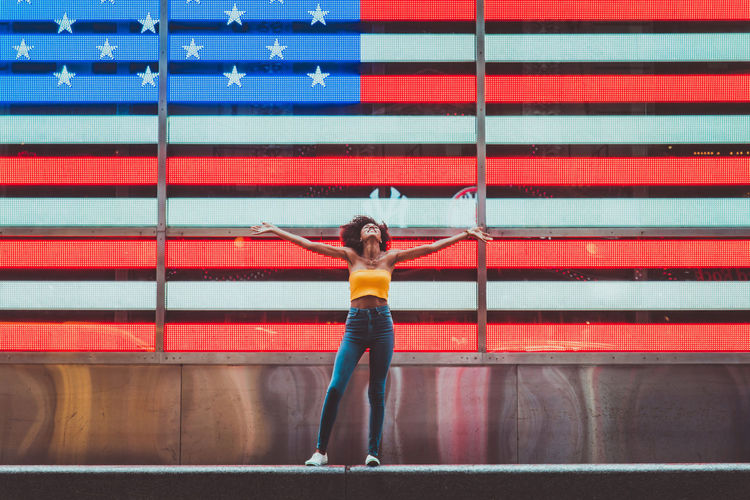 Full length of young woman standing with arms outstretched against neon american flag in city