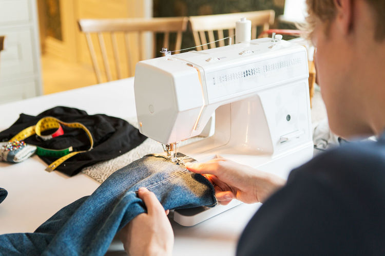 Cropped image of young man sewing denim jeans at home