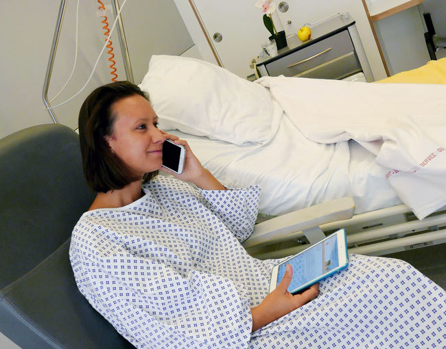 Young woman wearing hospital gown using mobile phone and digital tablet