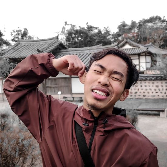 Young man pulling ear while standing against houses
