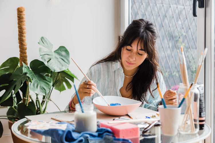 Concentrated talented female artist painting ceramic bowl while sitting at table with set of paintbrushes in light studio near door