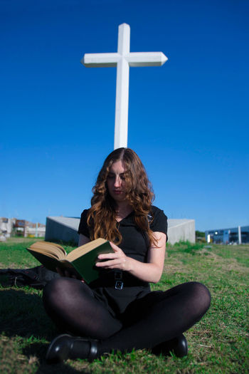 Young woman sitting on cross against blue sky