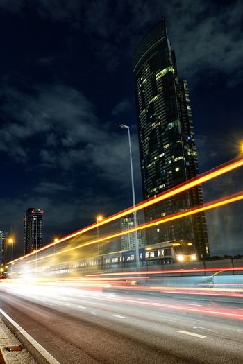 Light trails on road by illuminated buildings against sky at night