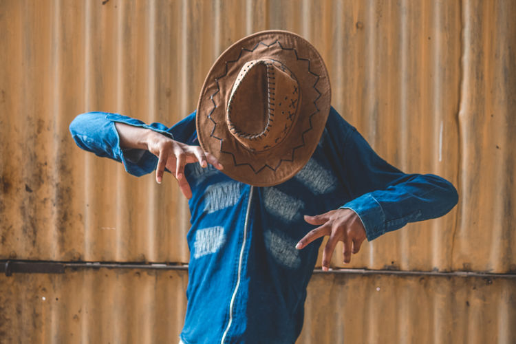 Teenage boy covering face with cowboy hat while standing against iron wall