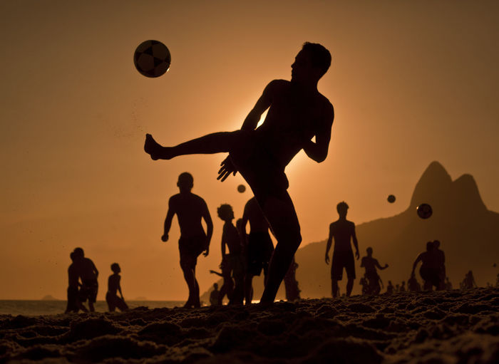 Silhouette friends playing soccer at beach against orange sky during sunset
