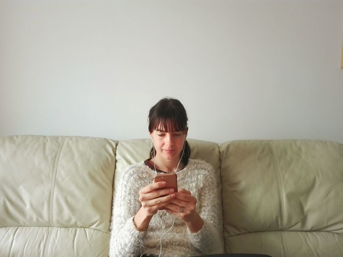 Woman listening music through smart phone while sitting on sofa against wall at home