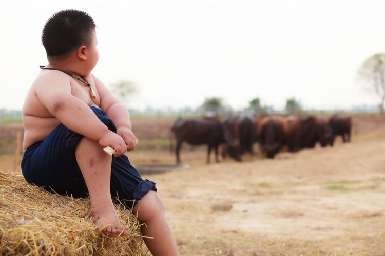 Shirtless boy sitting on hay bale against sky