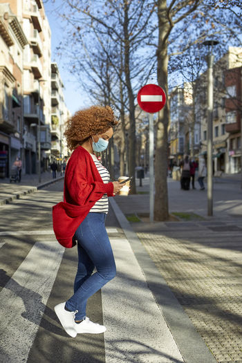 Afro woman wearing protective face mask using mobile phone while walking on road in city