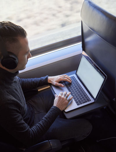 Side view of young male freelancer sitting on passenger seat in modern train and typing on netbook while working during trip
