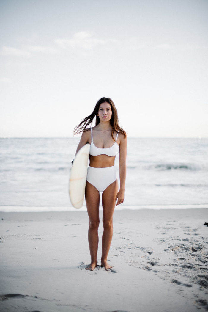 Full length portrait of young woman wearing bikini holding surfboard while standing at beach