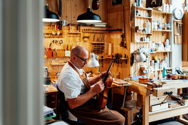 Side view of mature male luthier in apron and glasses sitting on chair and holding restored violin while working in workshop