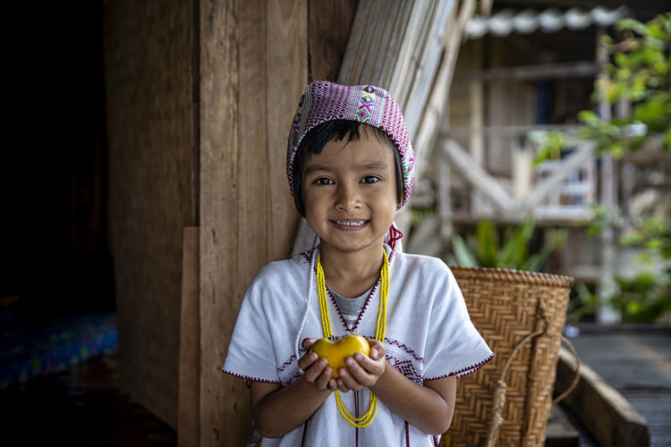 Portrait of smiling girl holding fruit while standing against wooden hut