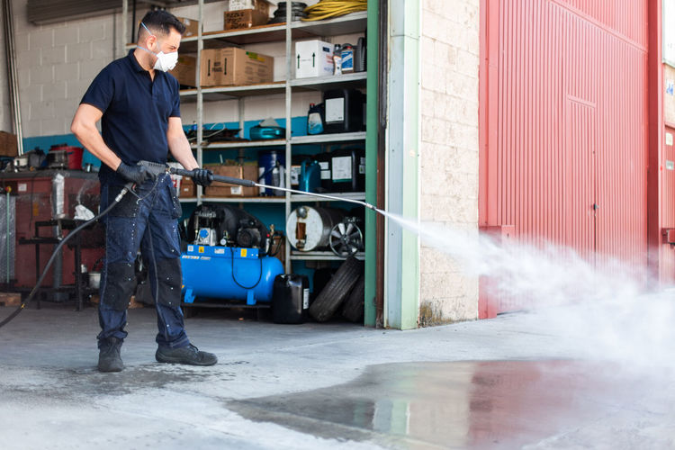 Full body man in respirator washing concrete ground near workshop while working in garage