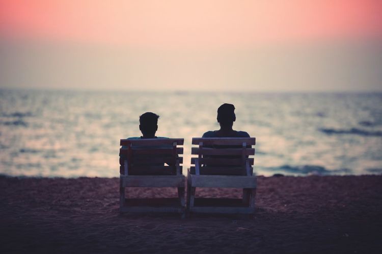 Rear view of couple sitting on chairs at beach against sky during sunset