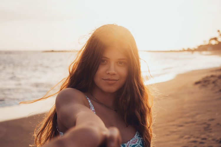 Portrait of young woman at beach against sky during sunset