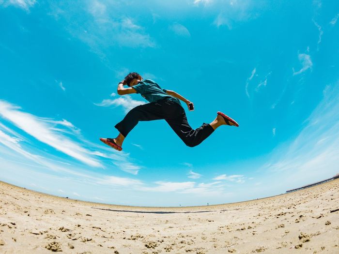 Low angle fish-eye lens shot of man jumping at beach against sky during sunny day