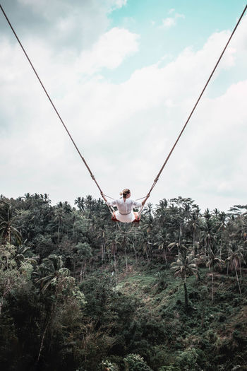 Woman sitting on swing by palm trees against sky