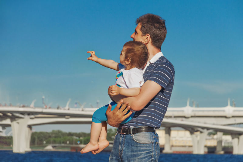 Dad joyfully clings to his daughter two years  against blue sky child shows with right hand to left