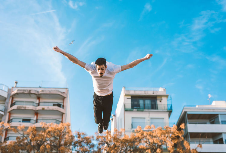 Full length of man with arms raised jumping against buildings and sky
