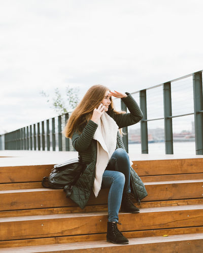 Side view of woman talking over smart phone while sitting on steps against sky