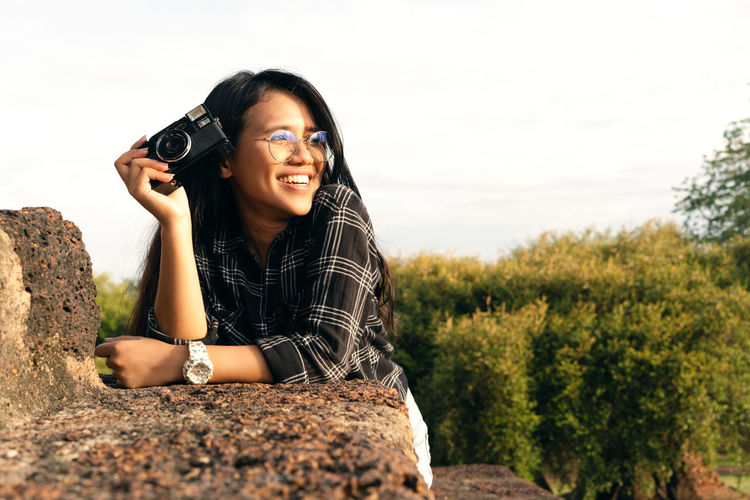 Portrait of smiling young woman photographing against sky
