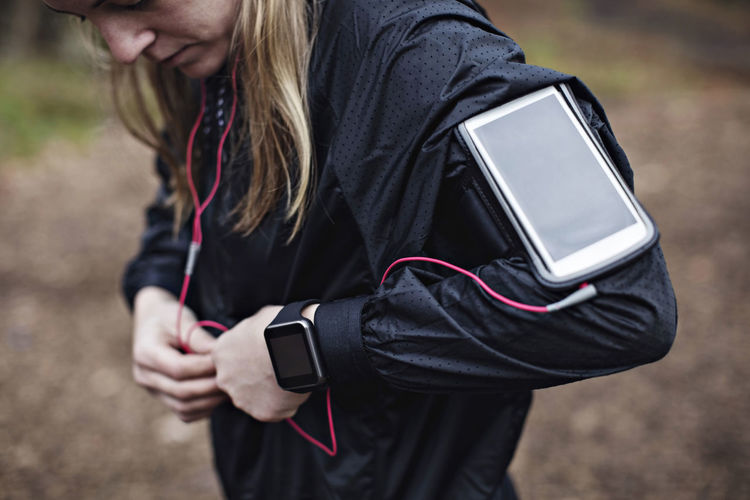 Midsection of female athlete with smart phone in arm band fixing headphones to jacket in forest