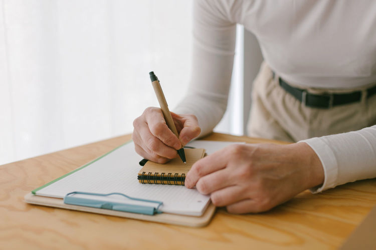 Faceless casual businesswoman bending on wooden table and writing in notepad with pen