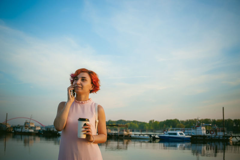 Mid adult woman talking on mobile phone while standing by river against sky during sunset
