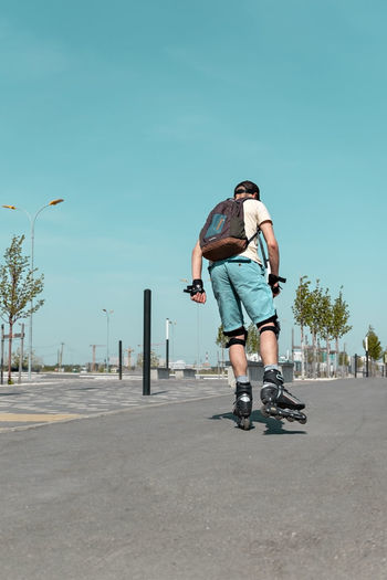 Rear view of young man with backpack in riding on roller skates on road in summer, roller blading