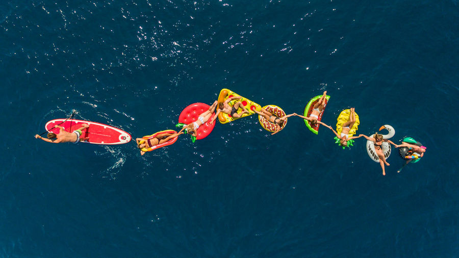 High angle aerial view of group of young travelers on colorful floaties holding hands in summer.