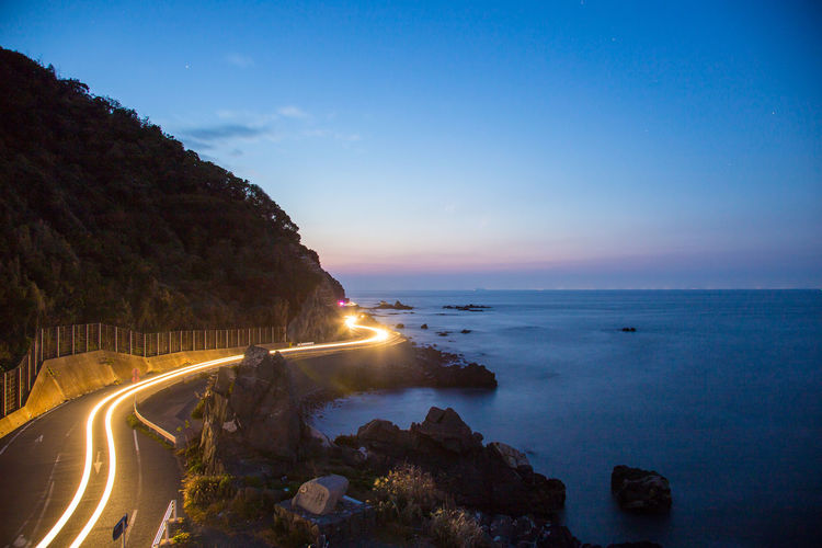 Light trail on road by sea against sky at dusk