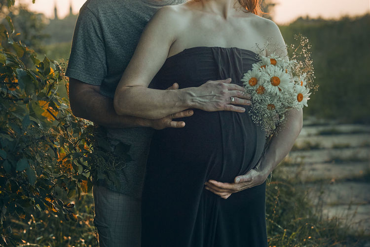 Pregnant woman and her husband hugging her tummy standing outdoors surrounded by nature. pregnancy