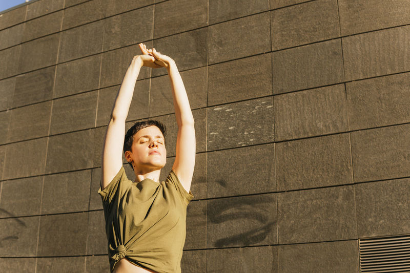 Woman stretching arms in front of wall on sunny day