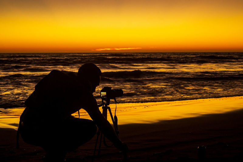 Silhouette man photographing at beach against sky during sunset