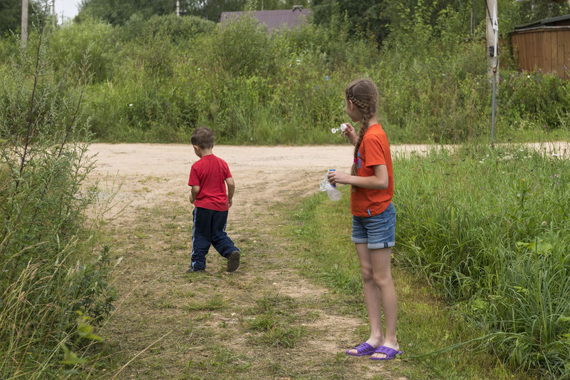 Girl blowing bubbles with boy walking on grass against plants