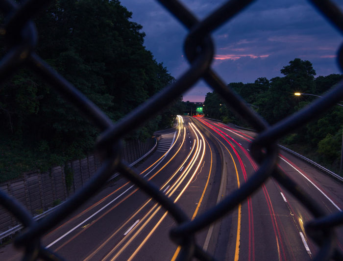 Light trails on road seen through chainlink fence at dusk