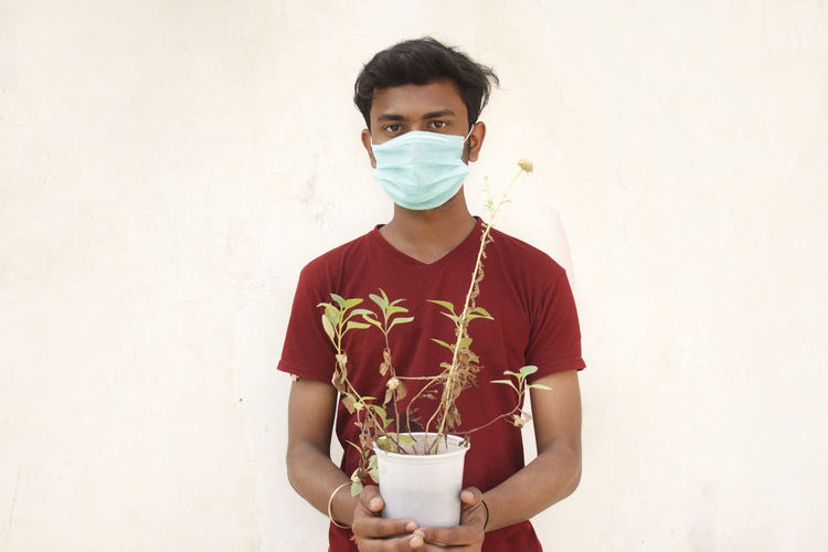 Portrait of young man wearing mask and holding plant standing against white background