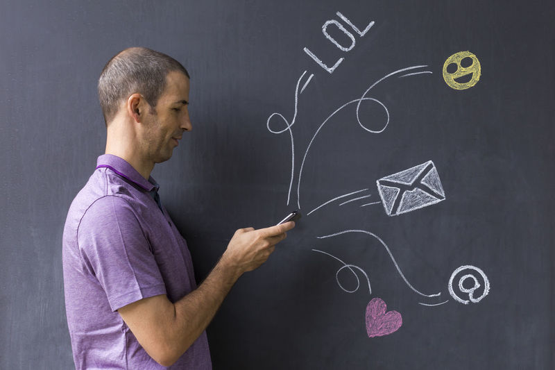 Side view man using mobile phone while standing by blackboard with various icons