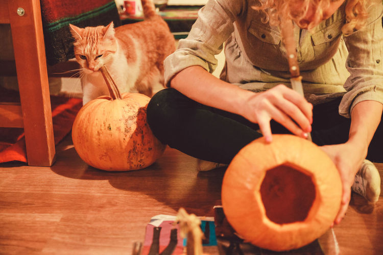 Woman sitting on floor while carving pumpkin lantern with cat at home