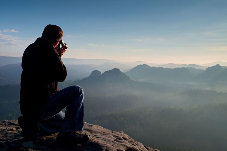 Dark hair man is taking photo by big mirror camera on the neck on the peak of mountains