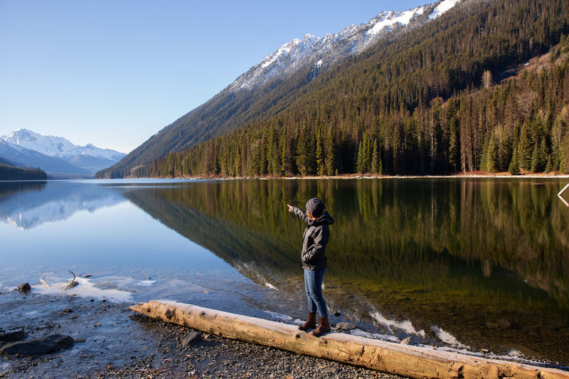 Side view of young woman walking on log at lakeshore against mountain