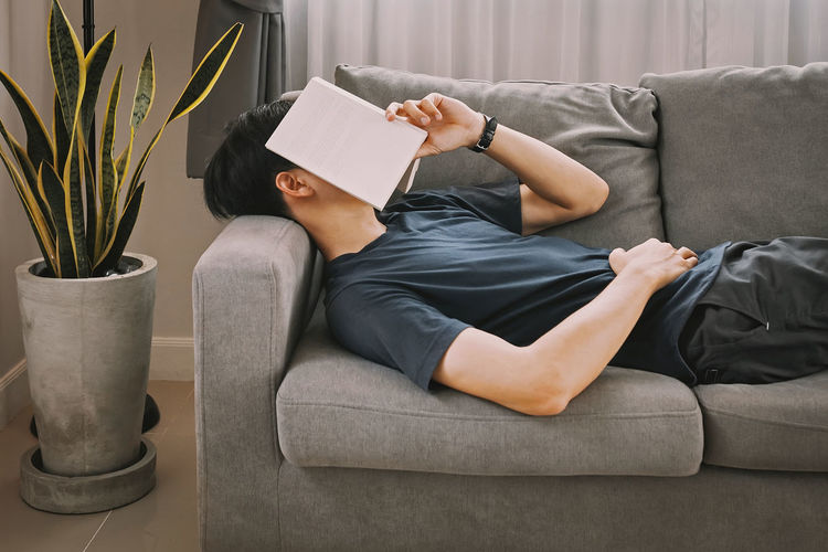 Man covering face with book while lying on sofa at home