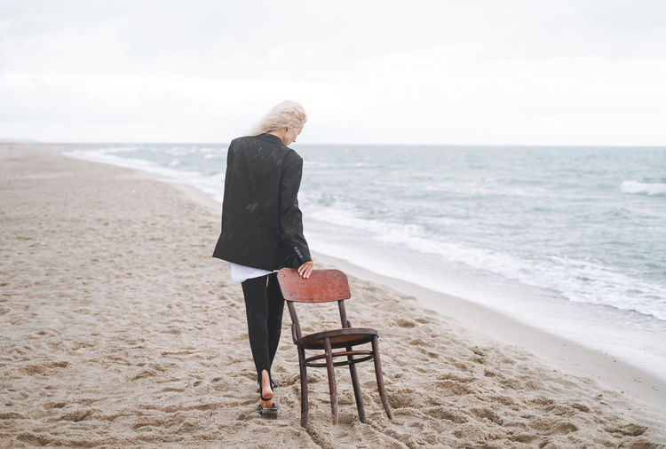 Portrait of elegant blonde woman in black suit with vintage wooden chair by sea in a storm