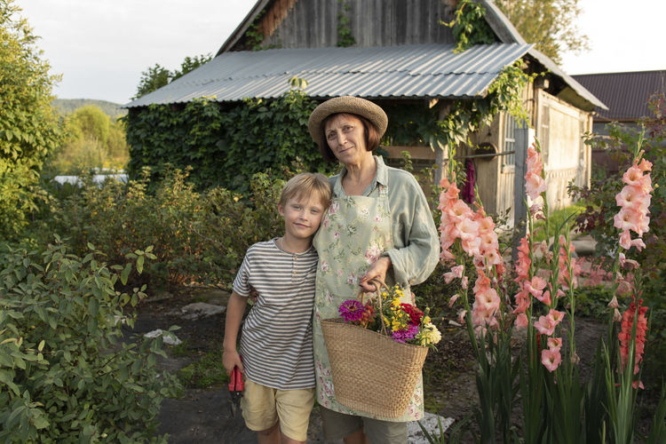 Smiling senior woman wearing hat standing with grandson in front of house