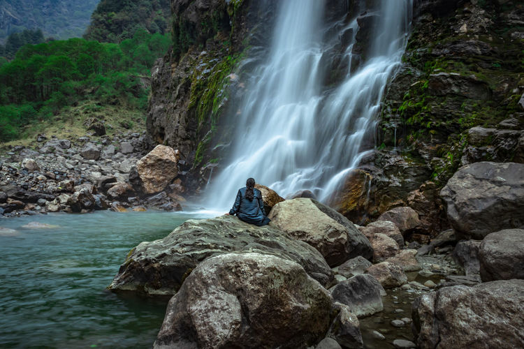 Girl sitting at rock in front of waterfall white water stream falling from mountains