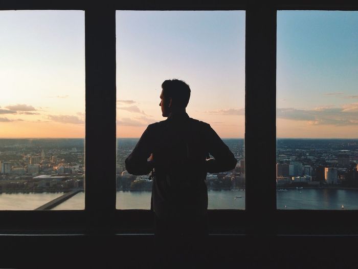 Rear view of man standing by window looking at city during sunset