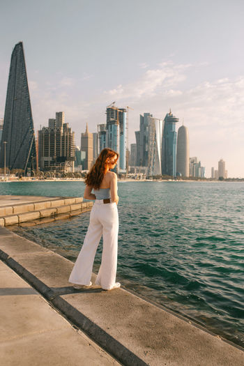 Full body young female tourist with long red hair standing on embankment and observing rippling sea water against downtown of doha, qatar