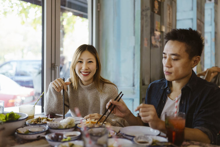 Portrait of smiling woman with chopsticks sitting with male friend having food at restaurant